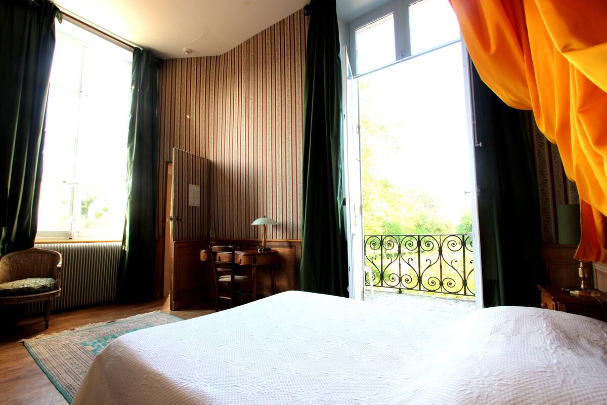 Une chambre d’hôtel avec un grand lit, un bureau avec une chaise, un radiateur et une vue sur la verdure par une fenêtre avec des rideaux.