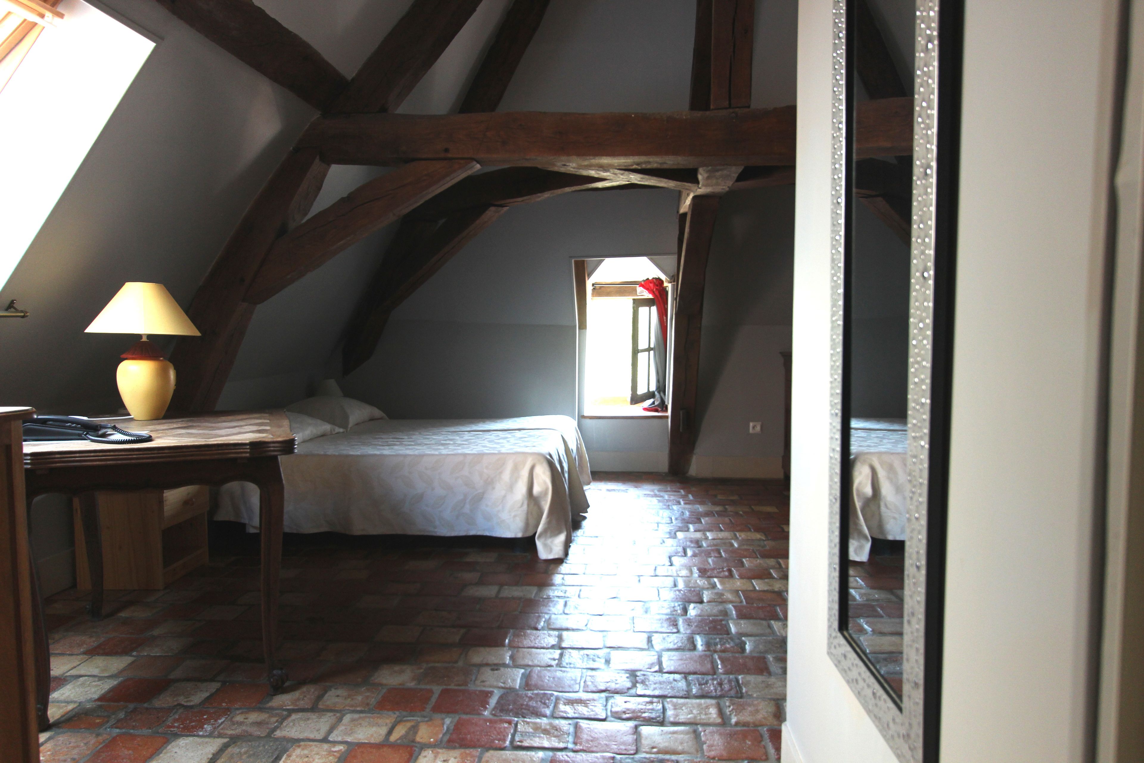 A bedroom with a brick floor, a bed, a desk with a lamp, and a window with a red curtain.