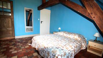 A bedroom with a brick floor, a bed with floral bedding, a wooden ceiling beam, a nightstand with a lamp, and a door with a mirror.