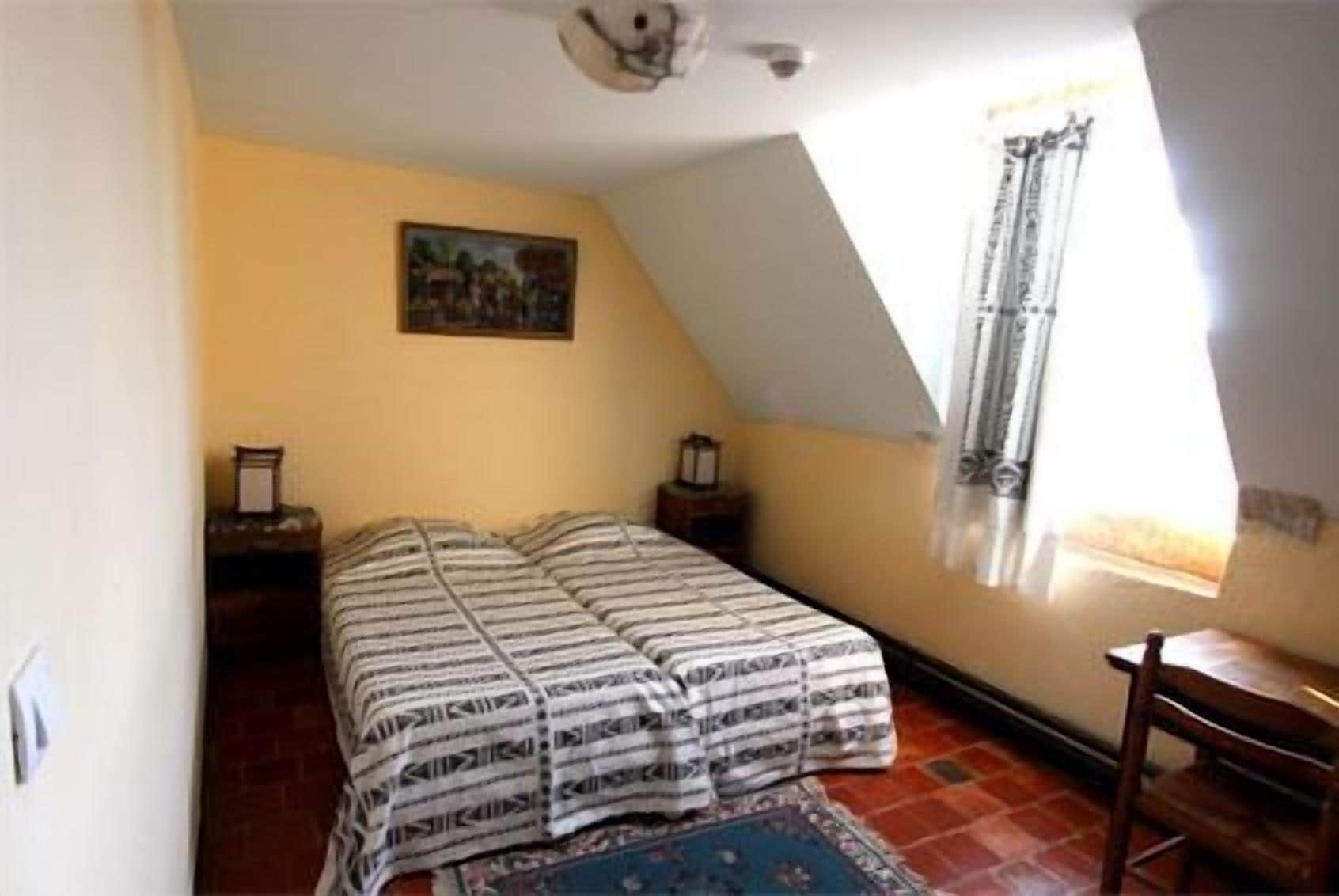 A small, cozy room with a sloped ceiling, a bed with striped bedding, a small table, and a window with curtains.