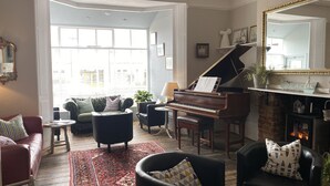 A spacious living room with a grand piano, comfortable seating, a large window, and a fireplace. - Marlborough Hotel (Shanklin)