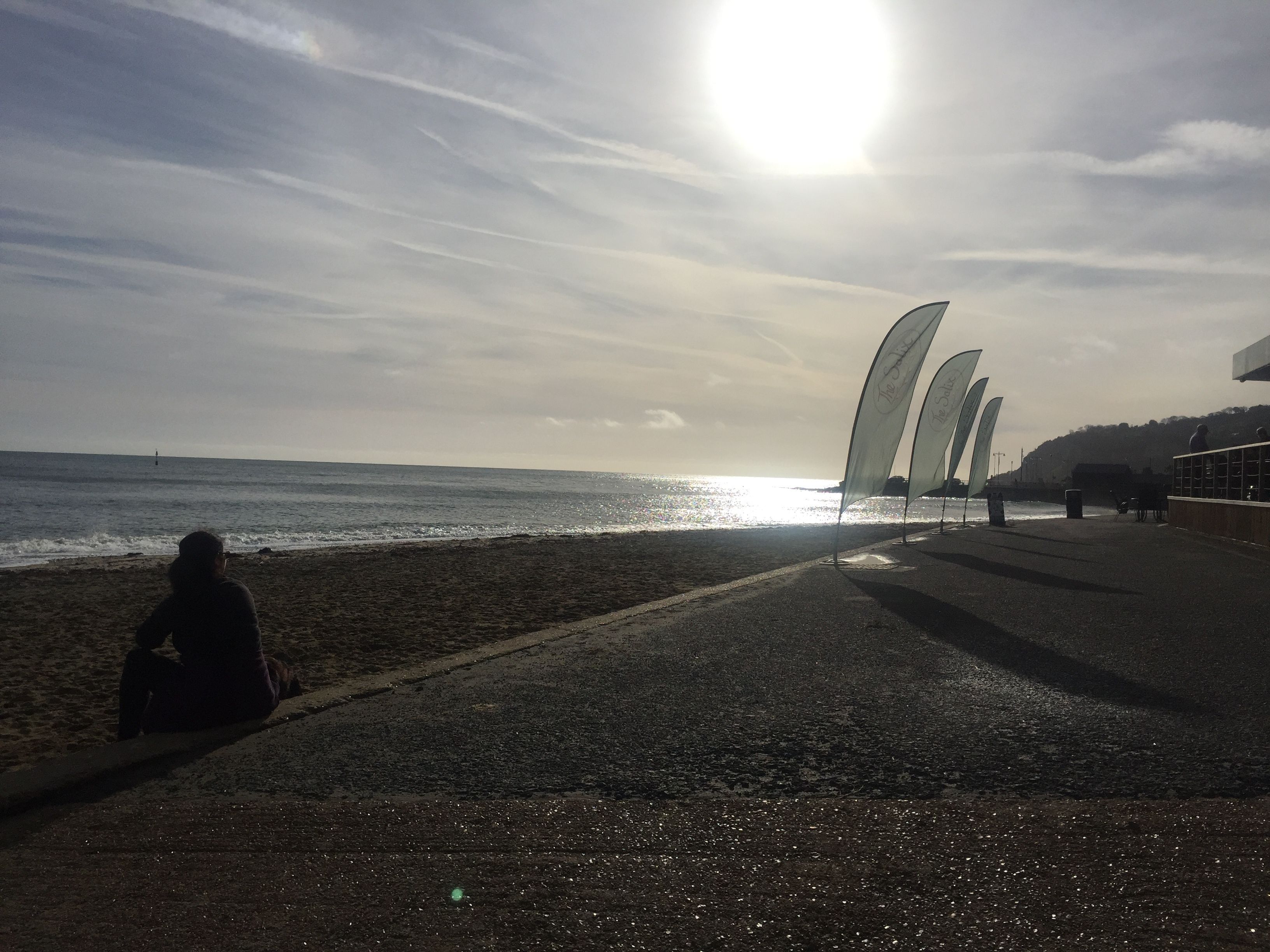 A beach with a person sitting on the sand, a clear sky, and a bright sun.