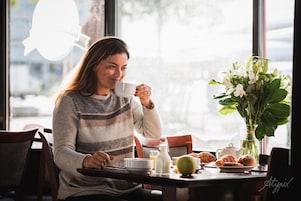 Una persona sentada a una mesa con una taza, un jarrón de flores y pasteles.