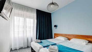 Quarto de hotel com duas camas, cabeceira de madeira, televisão de tela plana, luminária fixada na parede e uma grande janela com cortinas finas.