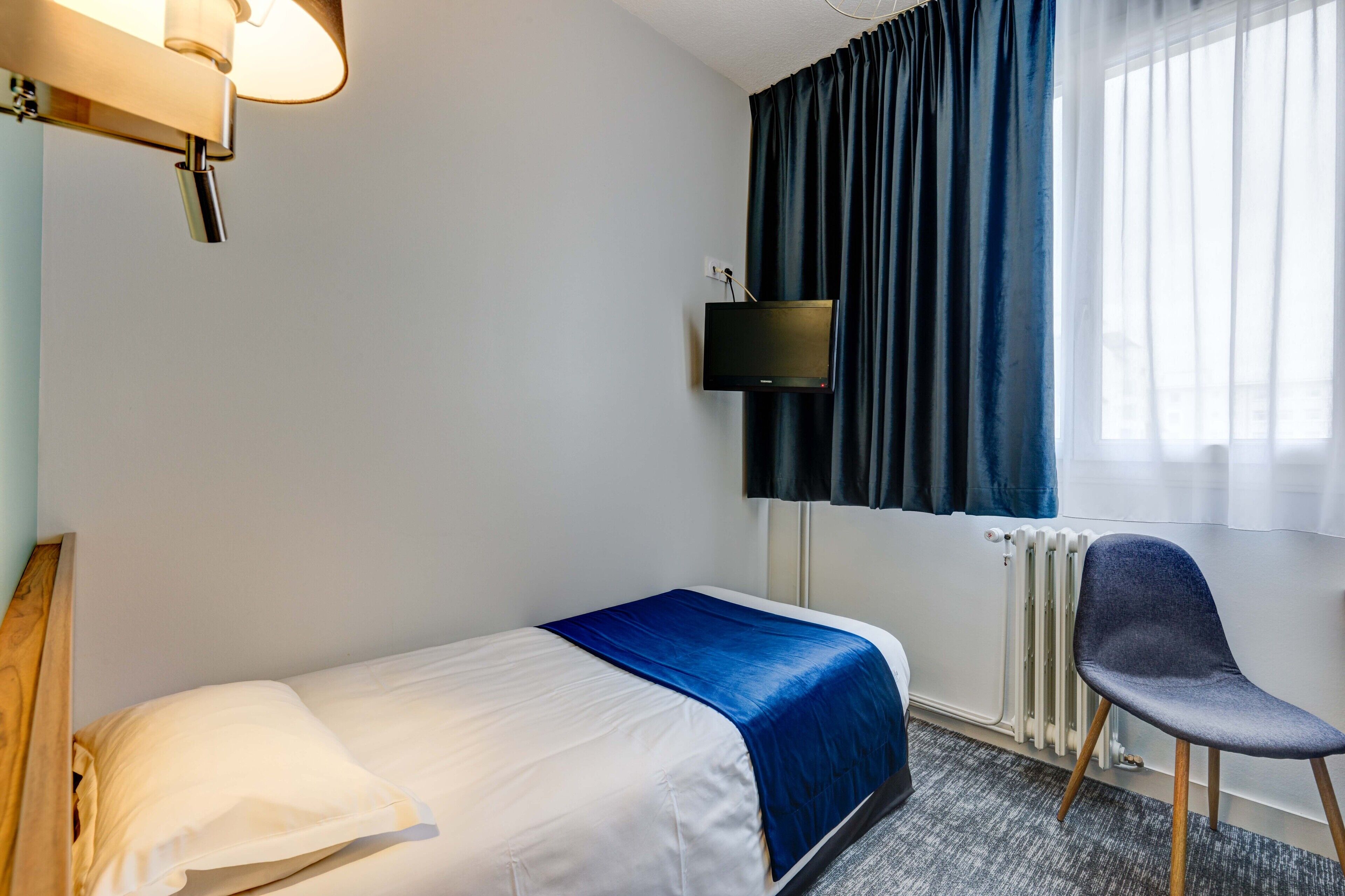 A hotel room with a single bed, a chair, a TV mounted on the wall, and a window with curtains.