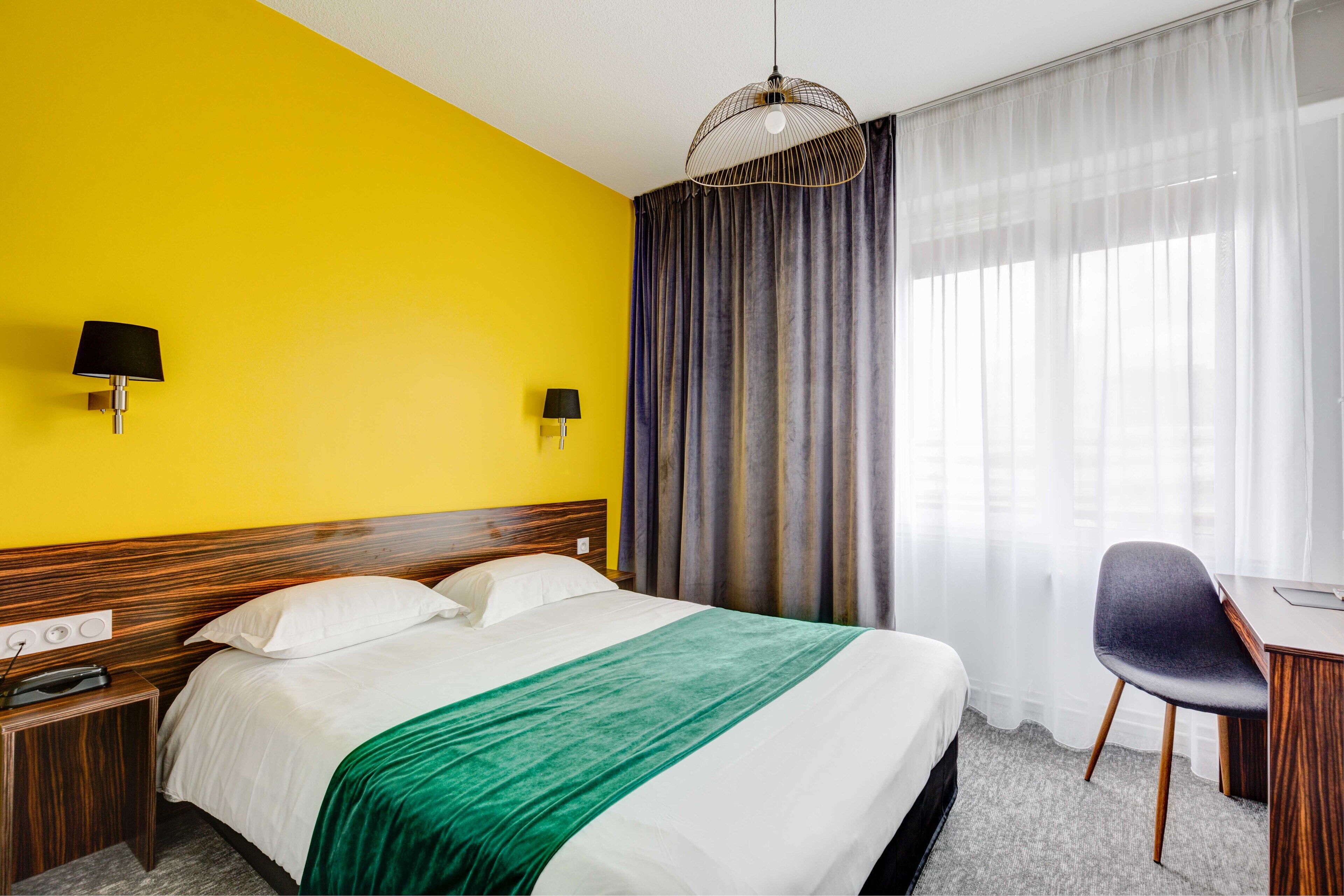 Una habitación de hotel con cama, escritorio, silla, lámpara y ventana con cortinas.