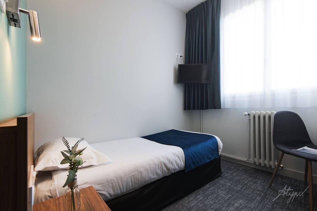 Habitación de hotel con una cama individual, un escritorio con silla, un televisor montado en la pared y una ventana con cortinas.