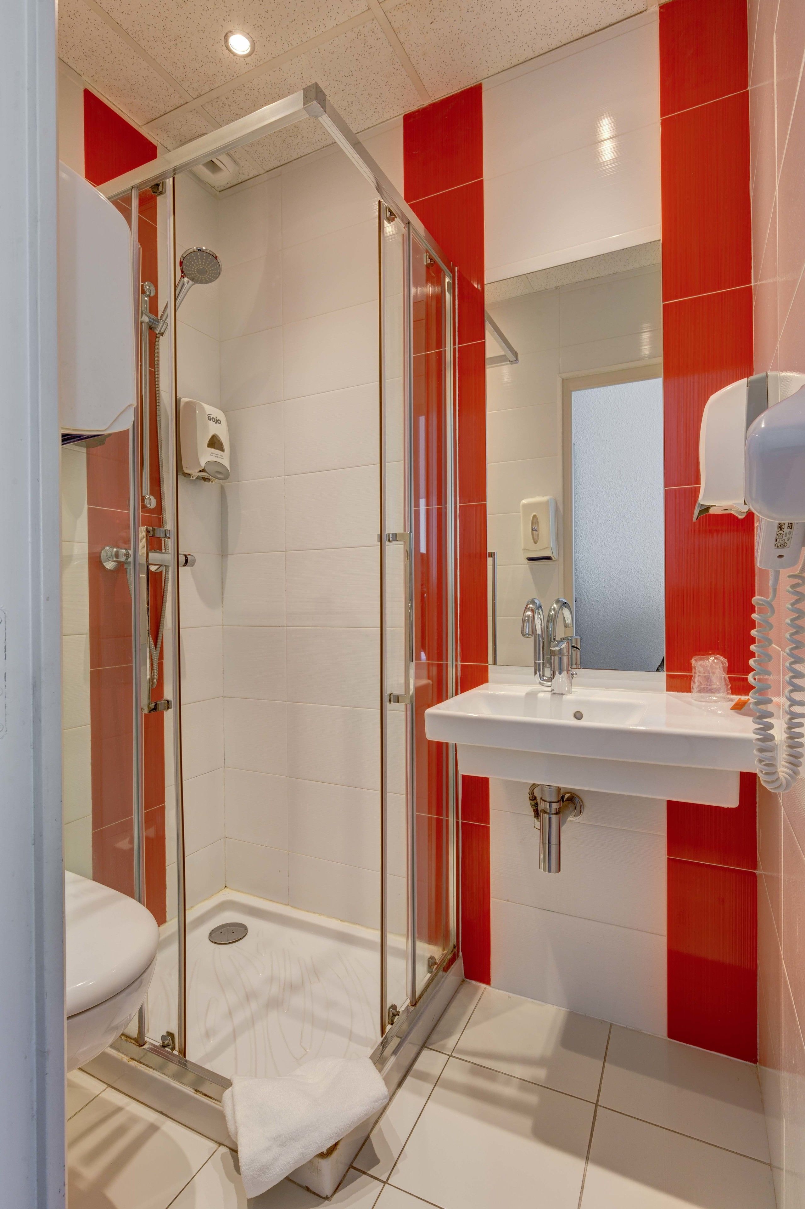 Un baño con ducha de vidrio, un lavamanos blanco con espejo encima y una pared de azulejos rojos.