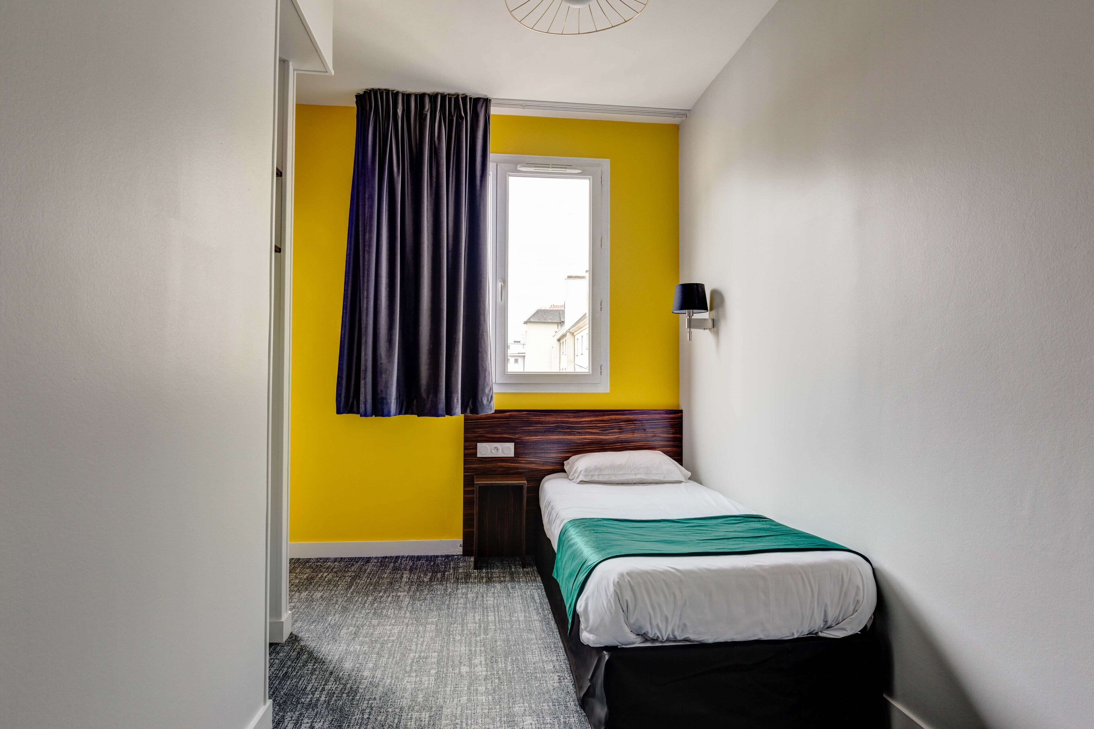 Una habitación de hotel pequeña y de colores vivos, con una cama individual, una ventana con cortinas, una lámpara de pared y vista a la calle.