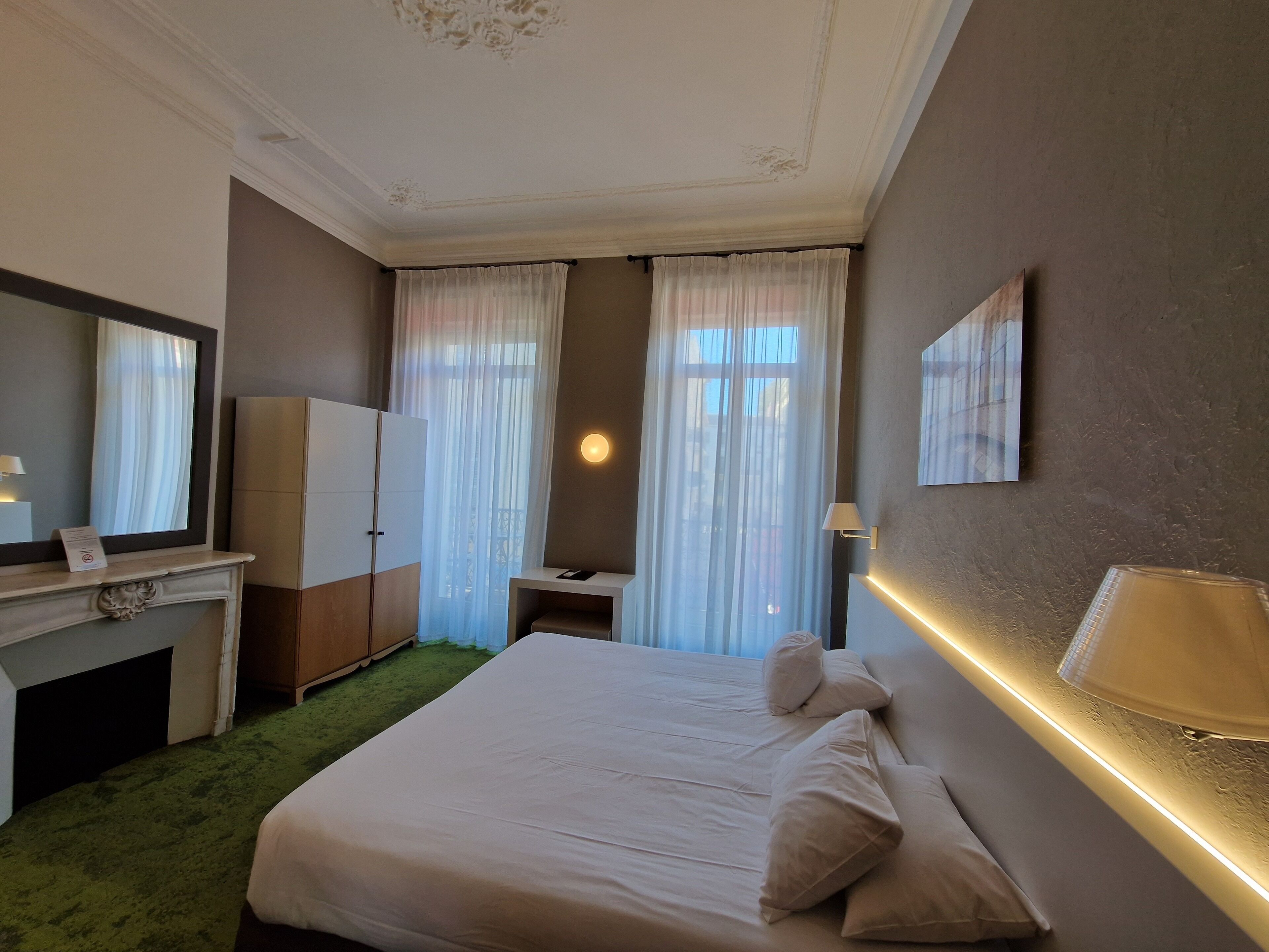 A hotel room with a large bed, a desk, a wardrobe, a fireplace, and a mirror.