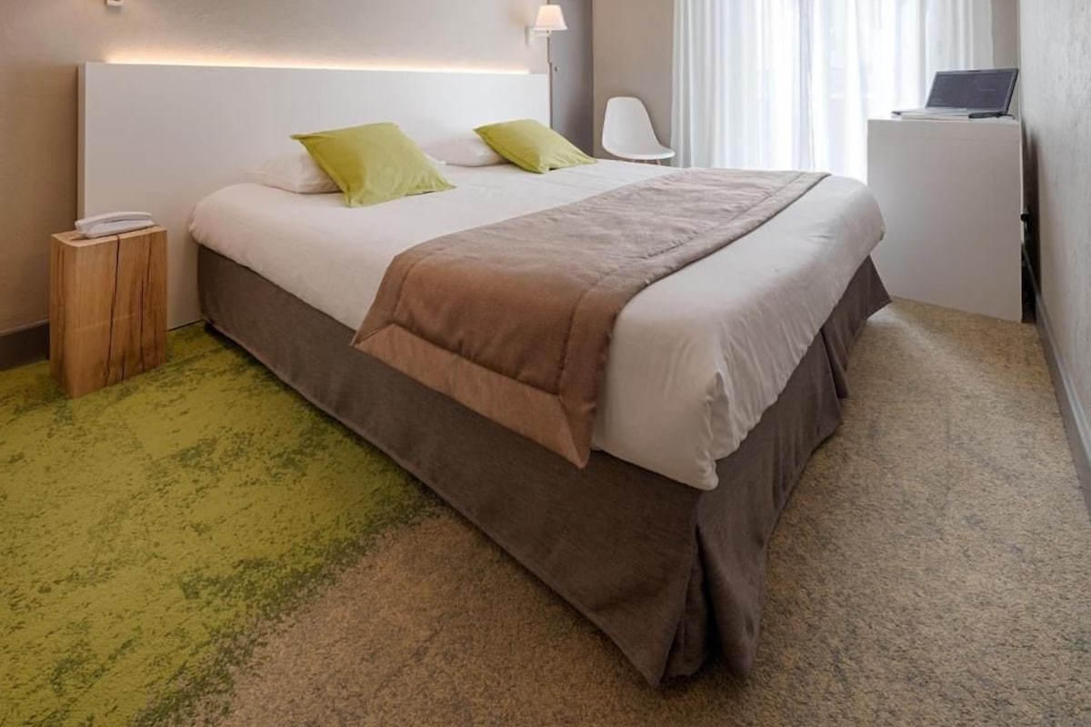 A hotel room with a large bed, two bedside lamps, a small wooden nightstand, a laptop on a desk, and sheer curtains.