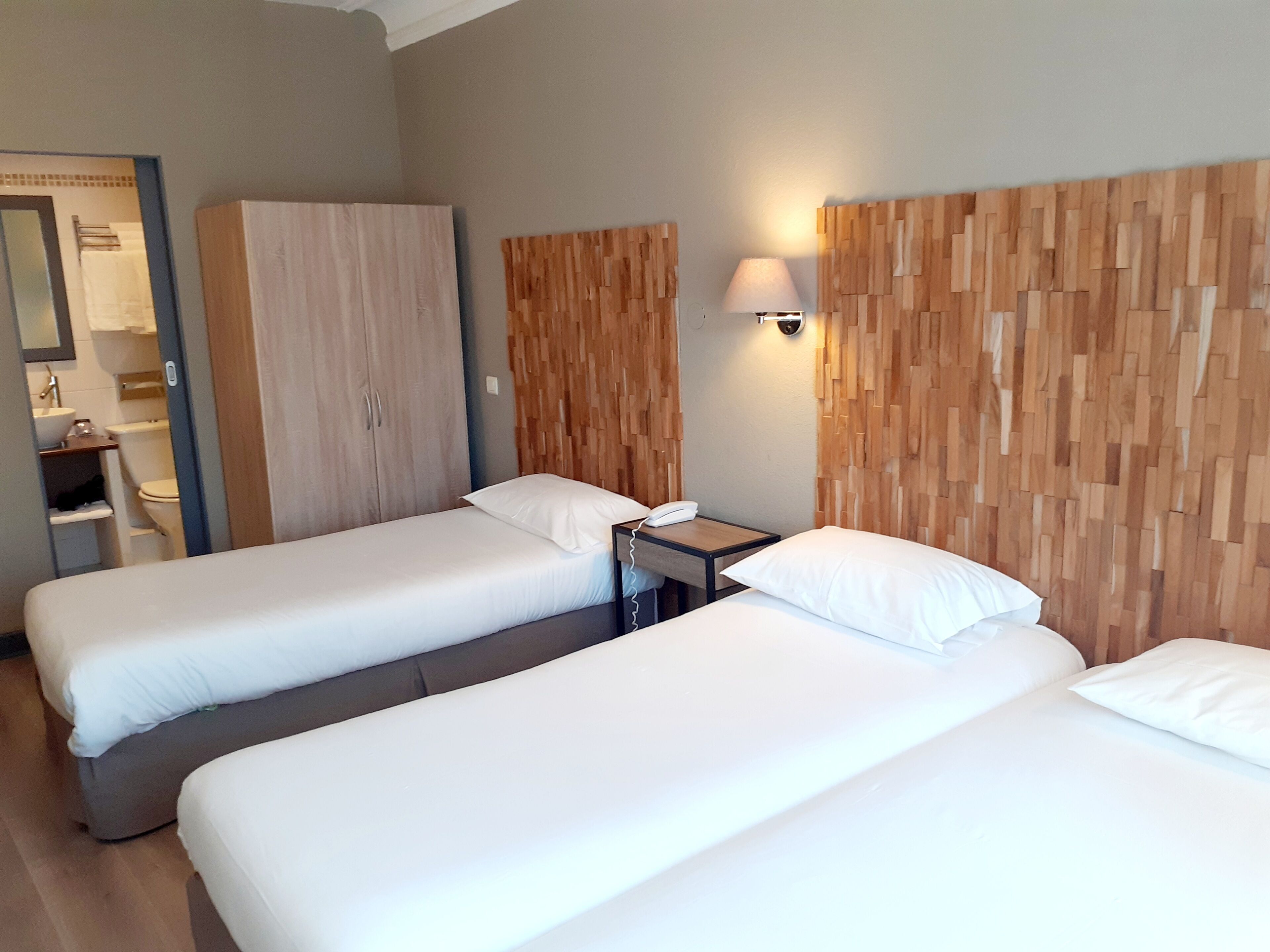 Une chambre d’hôtel avec deux lits simples, une tête de lit en bois, une table de chevet avec une lampe et une armoire.