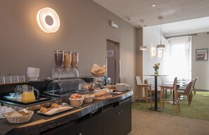 Um buffet de café da manhã com diversas opções de alimentos, um espaço para café e uma área de estar com mesas e cadeiras.