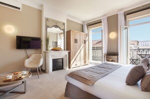 A modern hotel room with a large bed, a flat-screen TV, a fireplace, a balcony with a view of buildings, and a dining area with a tray of food.