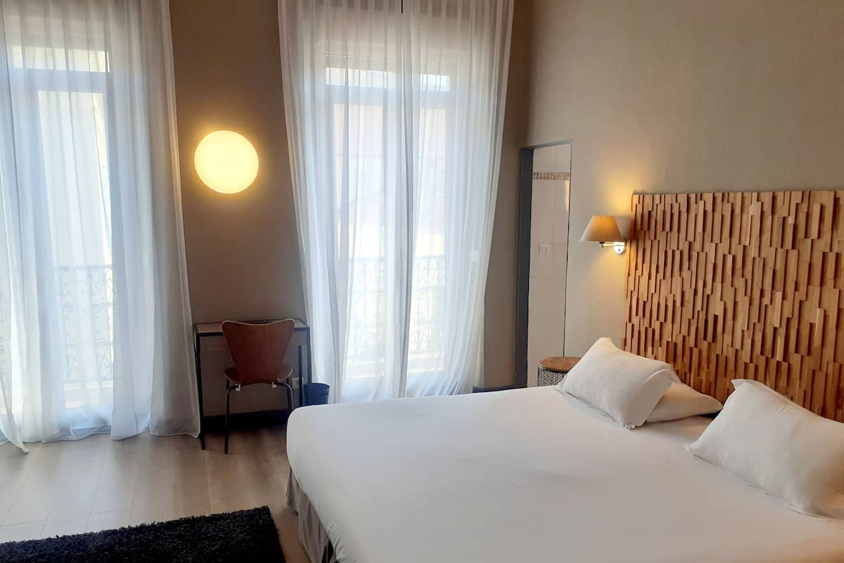 A hotel room with a large bed, a desk with a chair, and a wall-mounted lamp.