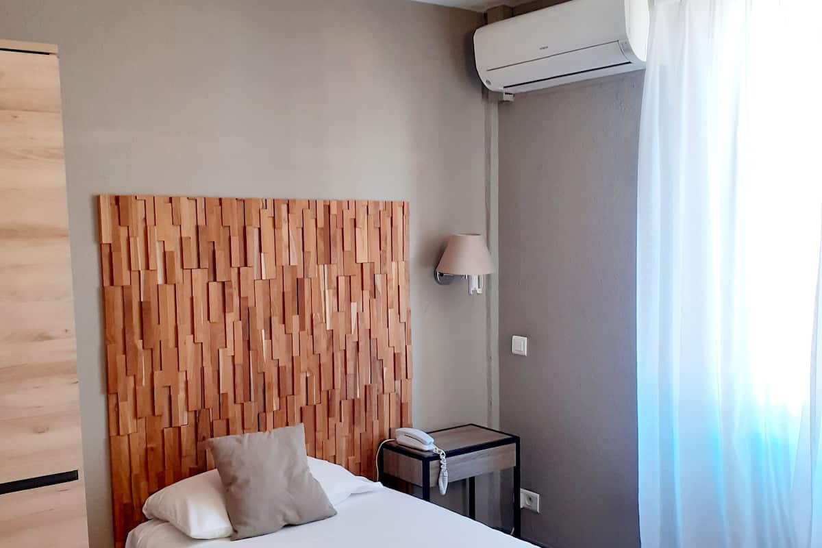 A hotel room with a wooden headboard, a bed with white linens, a bedside table with a lamp, and an air conditioning unit.