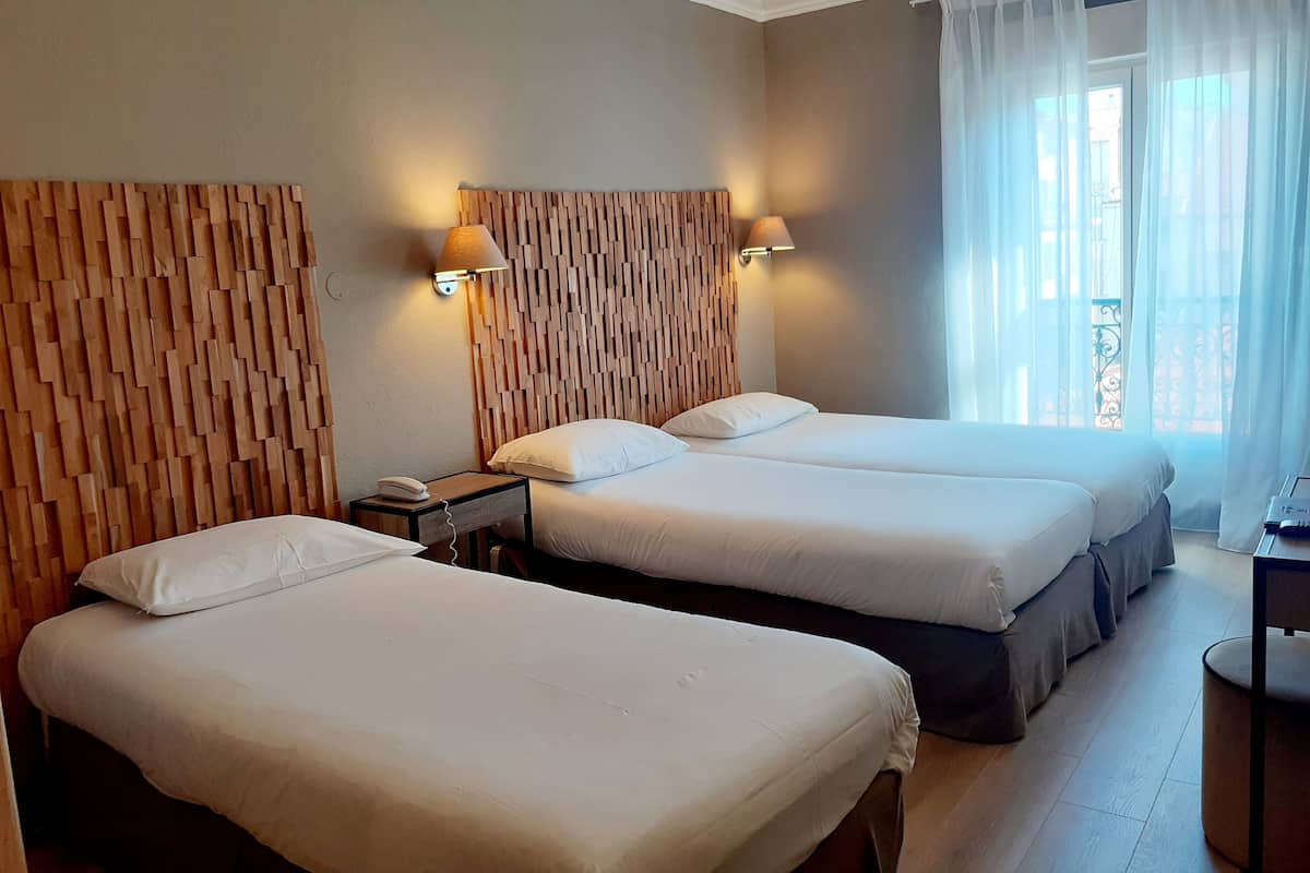 A hotel room with two beds, a wooden headboard, a bedside table with a lamp, and a window with sheer curtains.