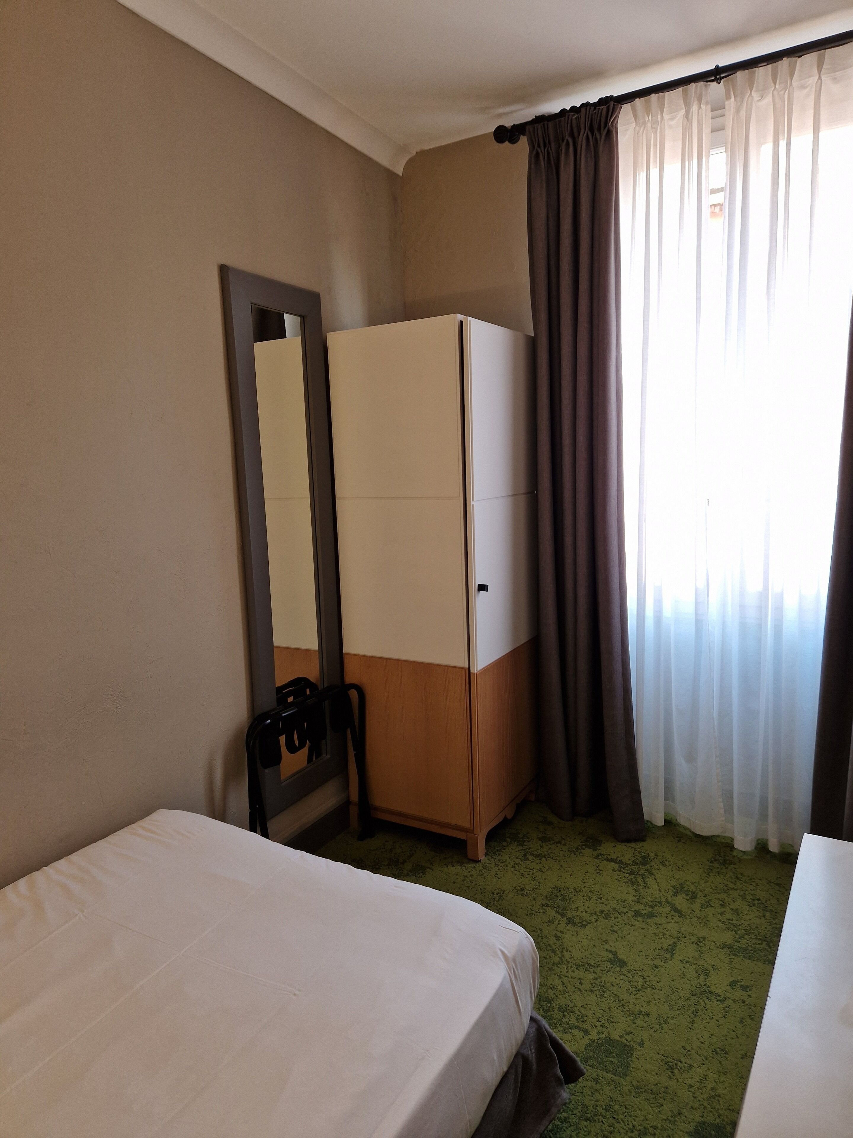 A hotel room with a bed, a wardrobe, a mirror, and a window with curtains.