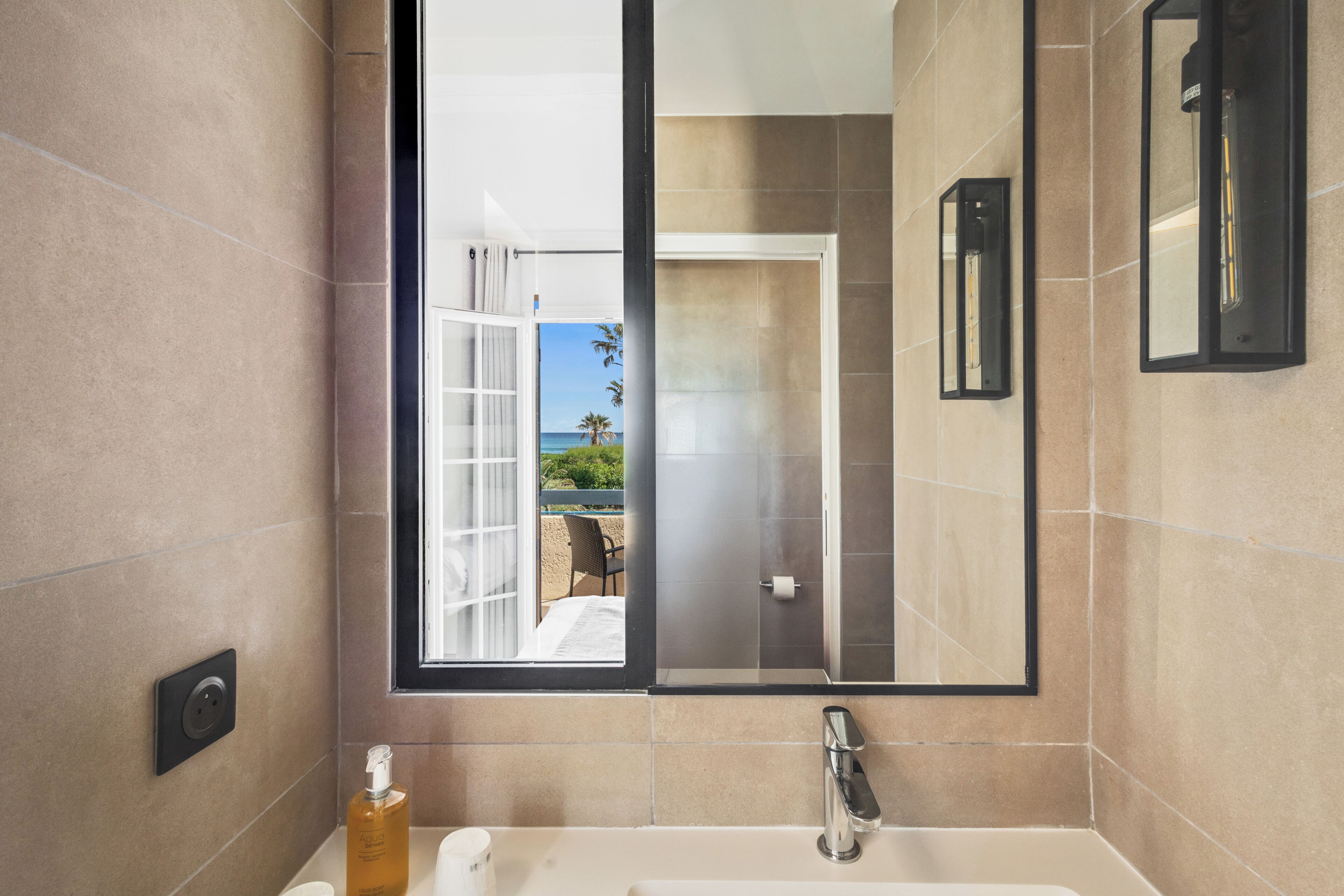 A bathroom with a large mirror, a sink, a towel dispenser, and a window offering a view of the beach.