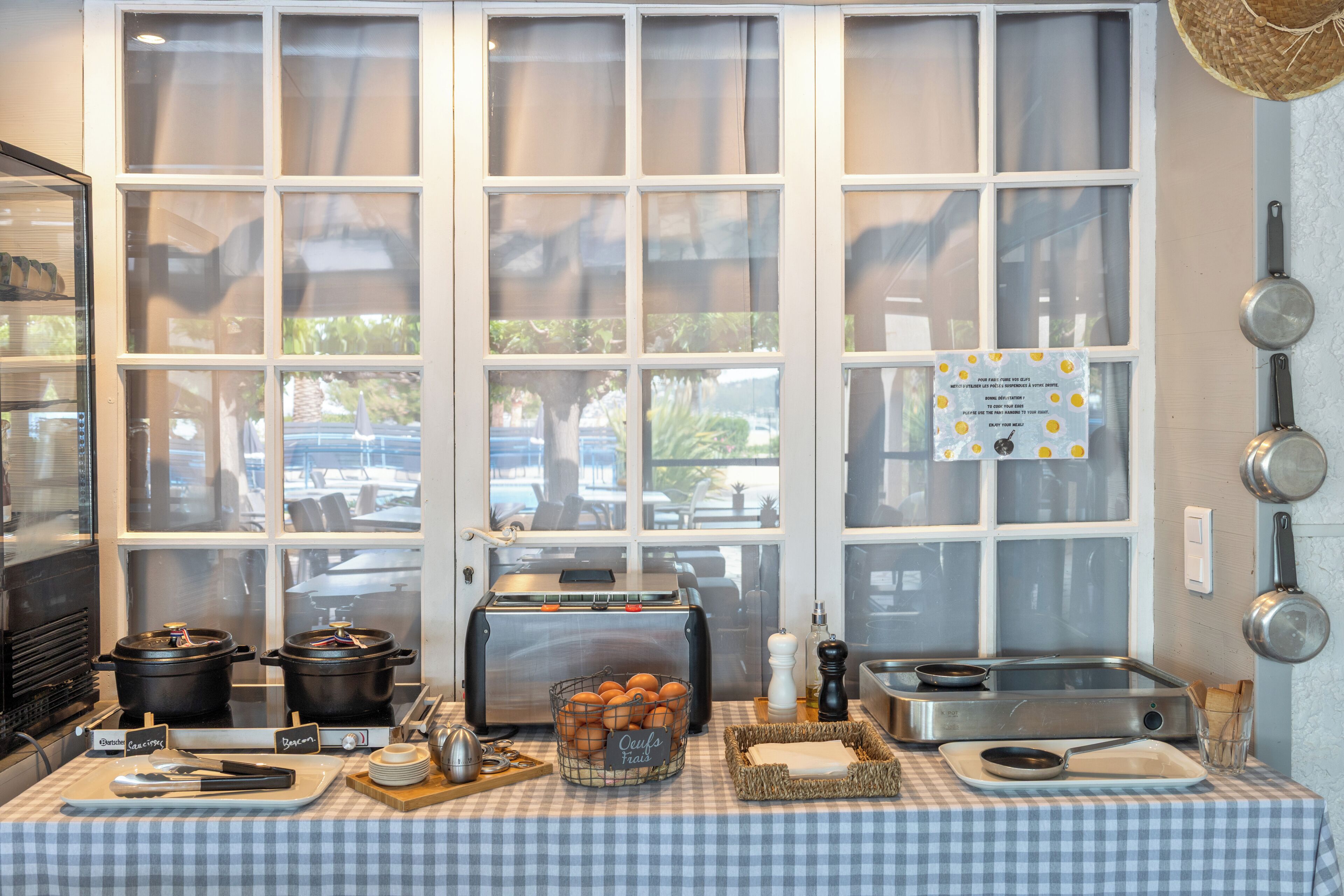 Una cocina con mantel a cuadros, máquina para pan, colgador para ollas y ventana con vistas al exterior.