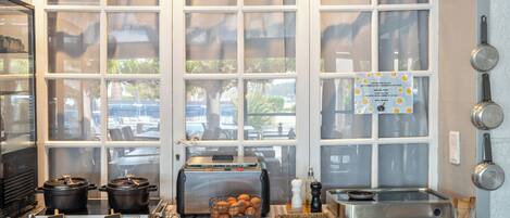 Una cocina con mantel a cuadros, máquina para pan, colgador para ollas y ventana con vistas al exterior.