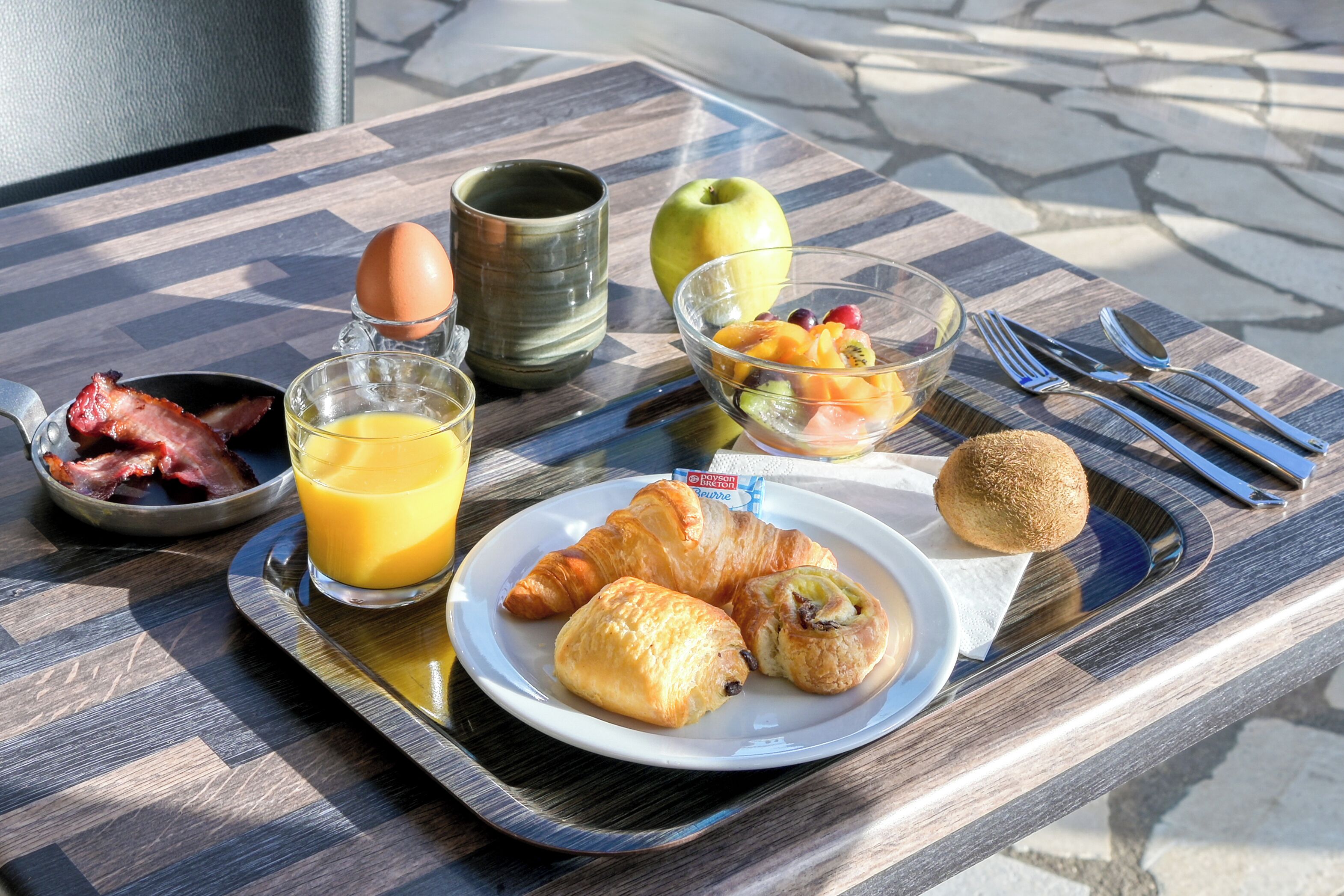 Bandeja de desayuno con cruasanes, tocino, huevos, jugo de naranja y fruta sobre una mesa al aire libre.
