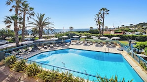 A swimming pool with lounge chairs and umbrellas, surrounded by palm trees and a view of the sea. - Hôtel La Nartelle (Sainte-Maxime)