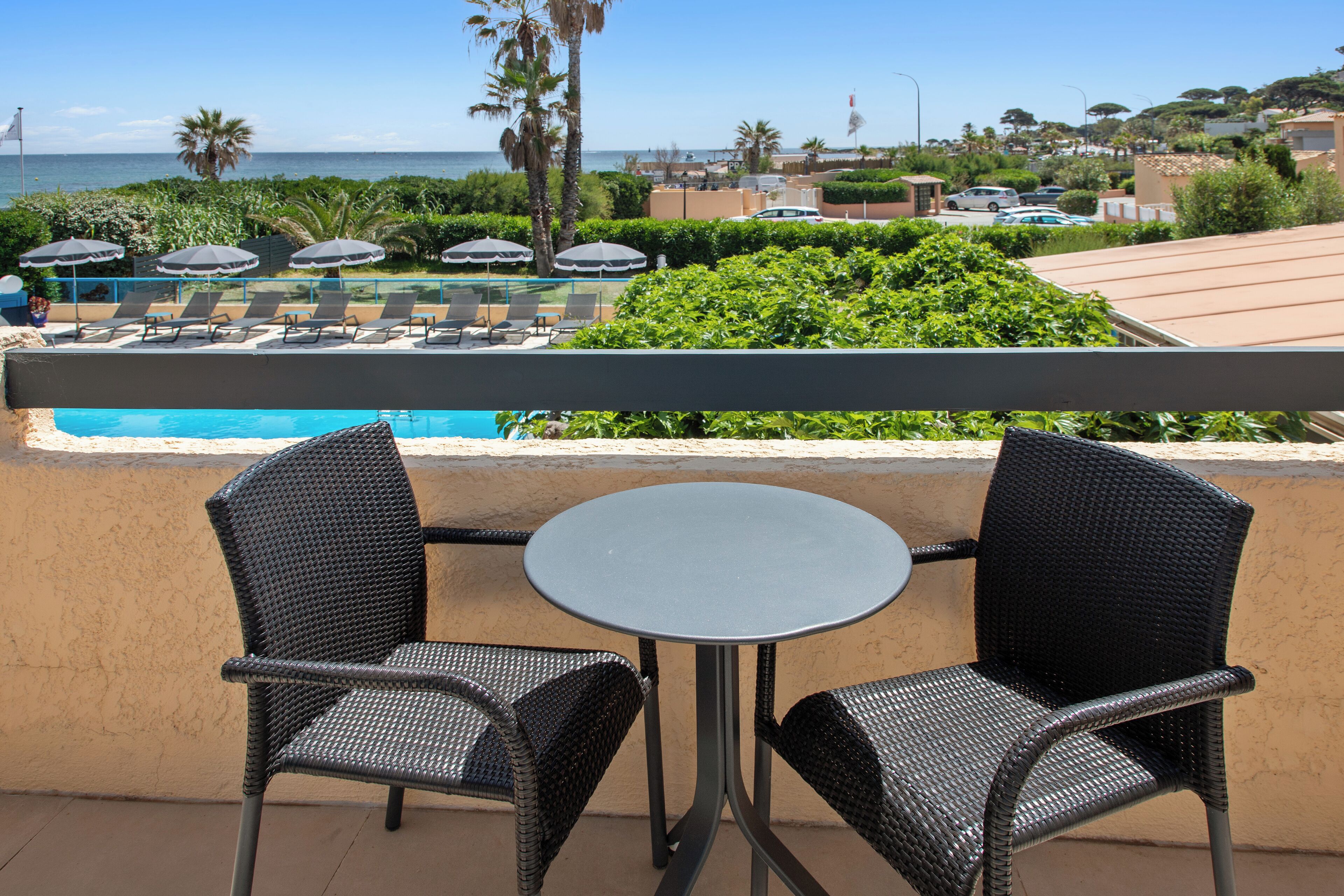 A balcony with two wicker chairs and a small round table, overlooking a pool and a beach.