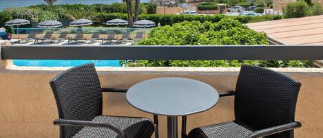 A balcony with two wicker chairs and a small round table, overlooking a pool and a beach.