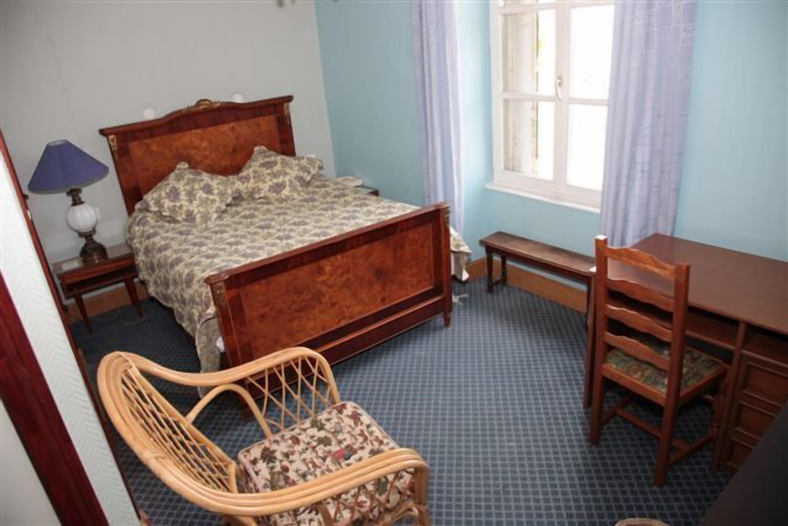 Une chambre à coucher avec un lit en bois, une chaise en osier, un bureau en bois et une fenêtre avec des rideaux.