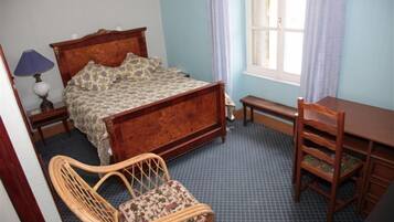 Une chambre à coucher avec un lit en bois, une chaise en osier, un bureau en bois et une fenêtre avec des rideaux.