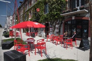 Un café al aire libre con sillas y mesas rojas, un paraguas rojo y un menú en una pizarra frente a un edificio de paredes de ladrillo y un balcón con flores.