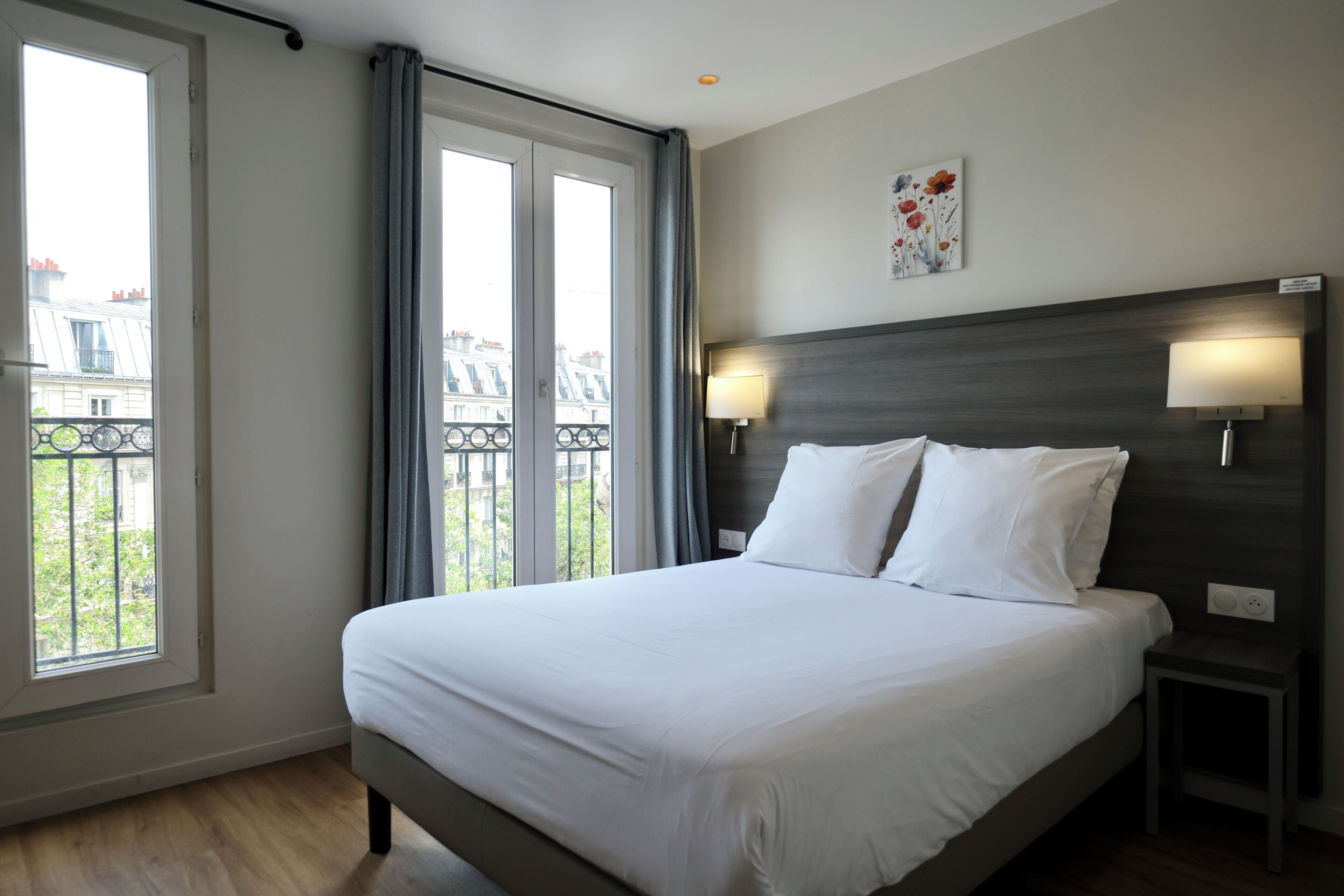 A hotel room with a large bed, two bedside lamps, a window with a view of buildings, and a small table.