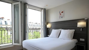 Una habitación de hotel con una cama grande, dos lámparas de noche, un ventanal con vistas a edificios y una mesita pequeña.
