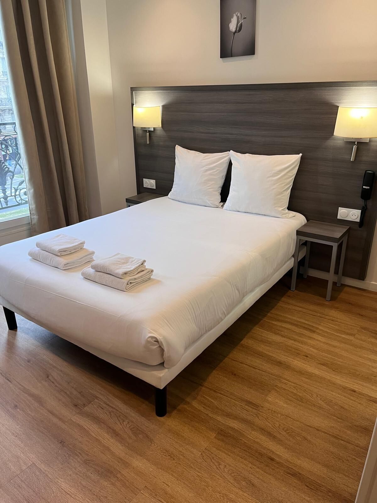 Una cama bien hecha con sábanas y toallas blancas, un cabecero de madera oscura, lámparas de noche y una mesita con un teléfono.