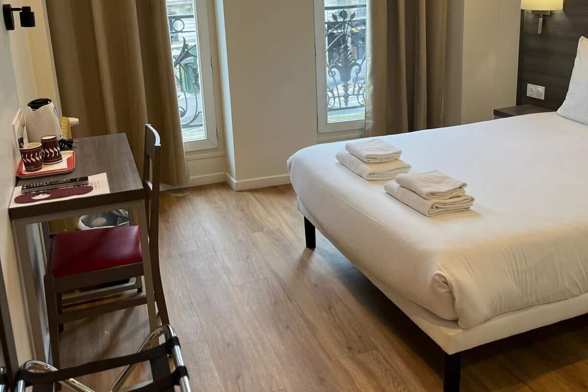 Een hotelkamer met een bed, een bureau met stoel, een raam met gordijnen en uitzicht op gebouwen.