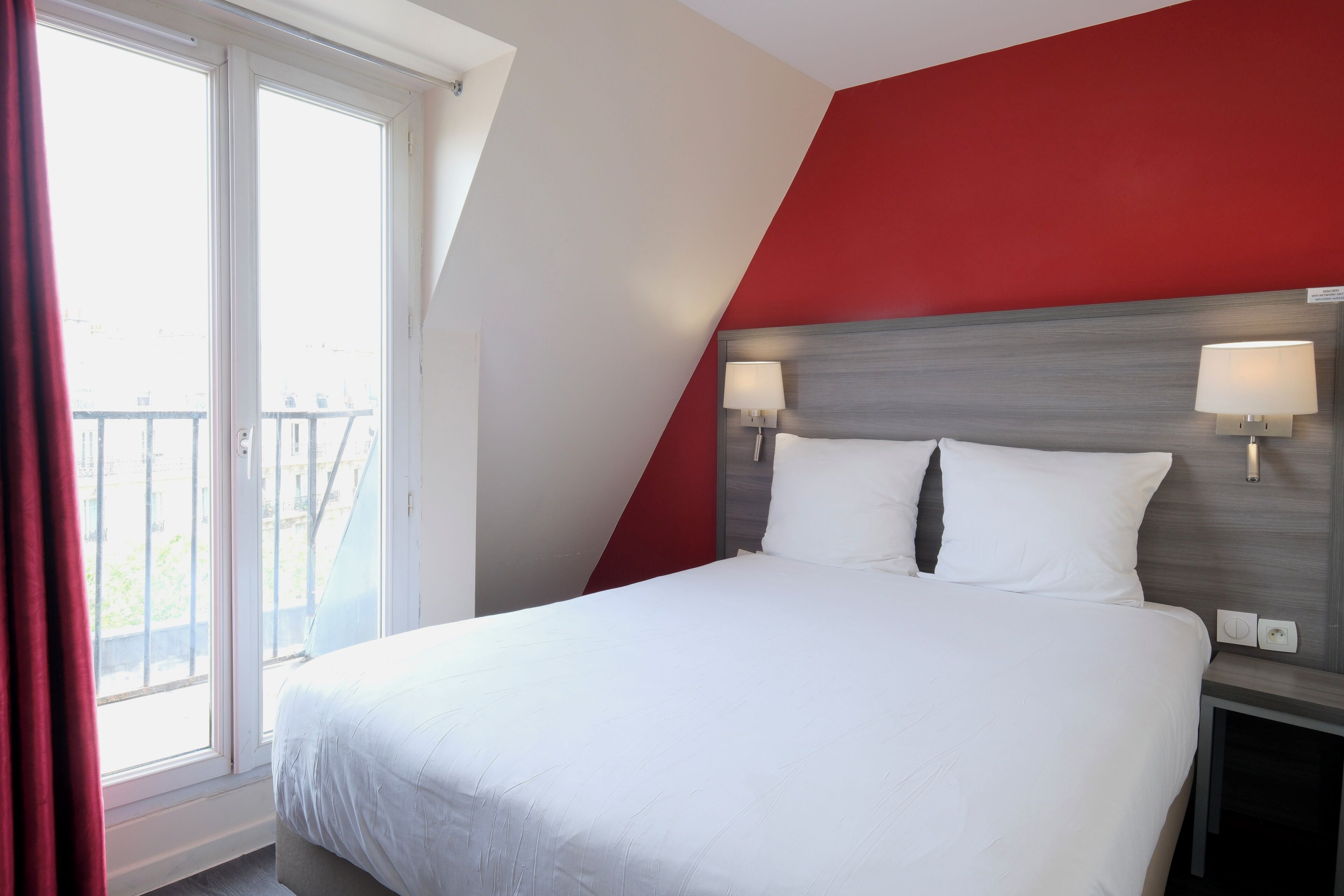 A hotel room with a bed, two bedside lamps, a red accent wall, and a window with curtains.