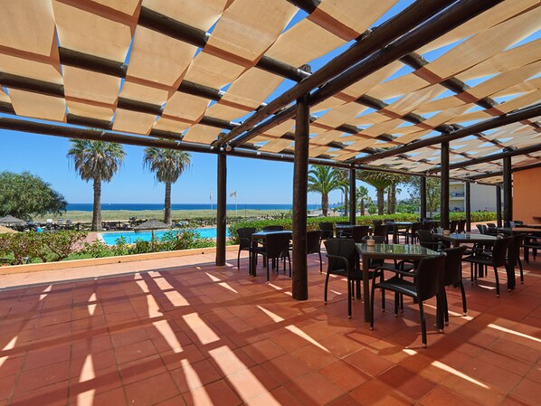 An outdoor patio with tables and chairs, overlooking a pool and the ocean. - Meia Praia Lagos - Beach Club Apartments (Lagos)