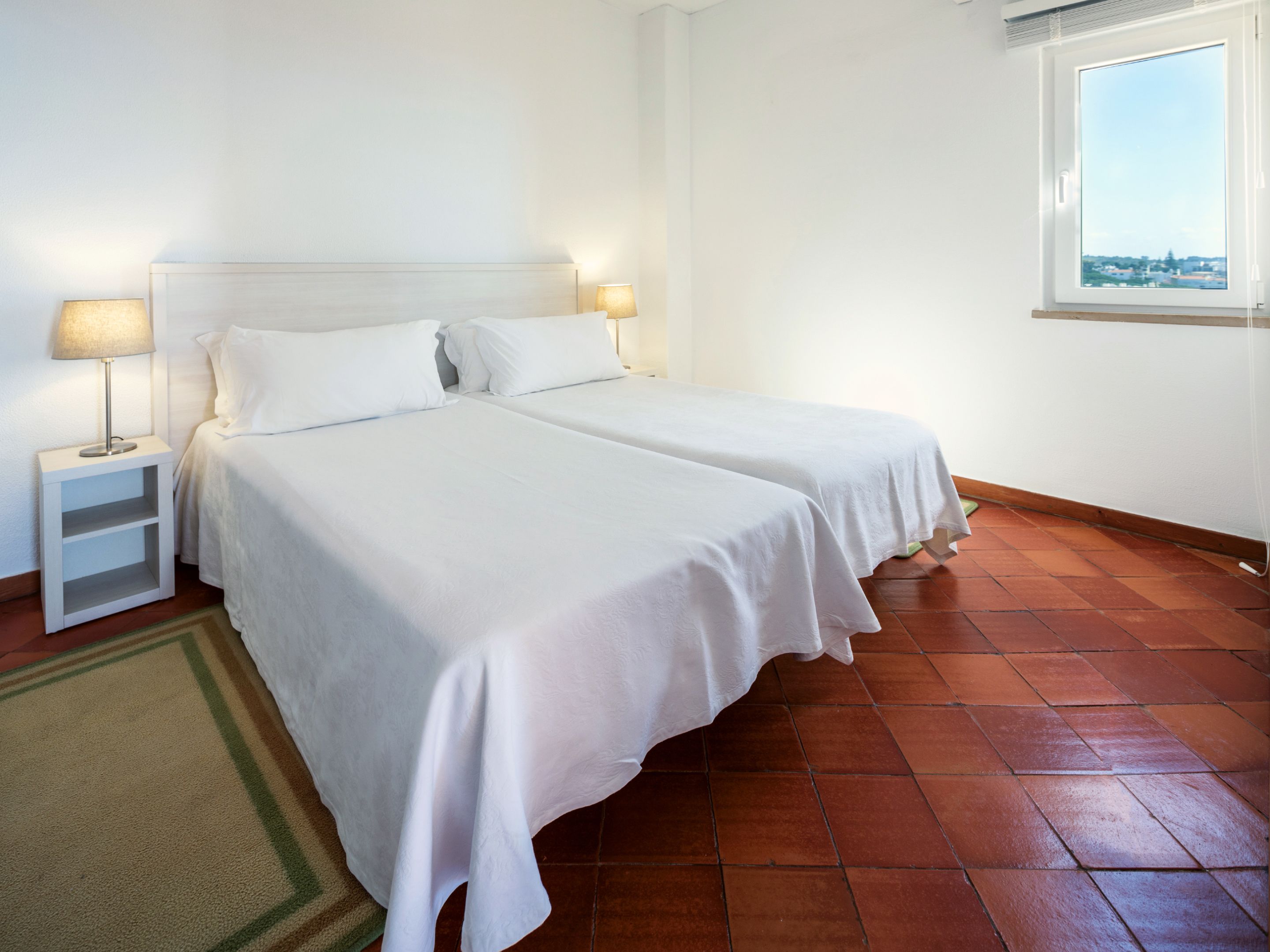 A room with two beds, a nightstand with a lamp, a window with a view, and a tiled floor.