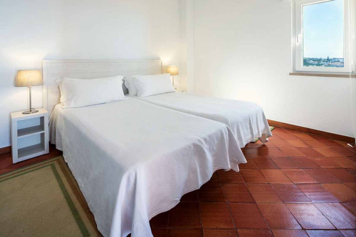 A room with two beds, a nightstand with a lamp, a window with a view, and a tiled floor.