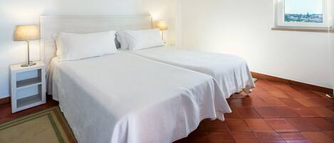 A room with two beds, a nightstand with a lamp, a window with a view, and a tiled floor.