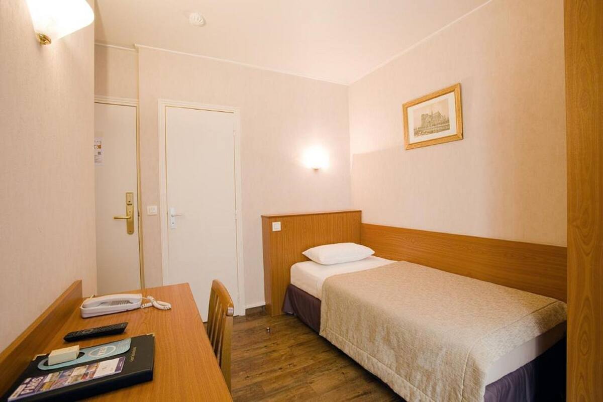 Une petite chambre propre, avec un lit simple, un bureau muni d’un téléphone et de télécommandes, et une porte.