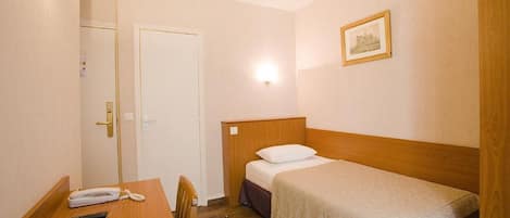 Una habitación pequeña y limpia con una cama individual, un escritorio con teléfono y controles remotos, y una puerta.
