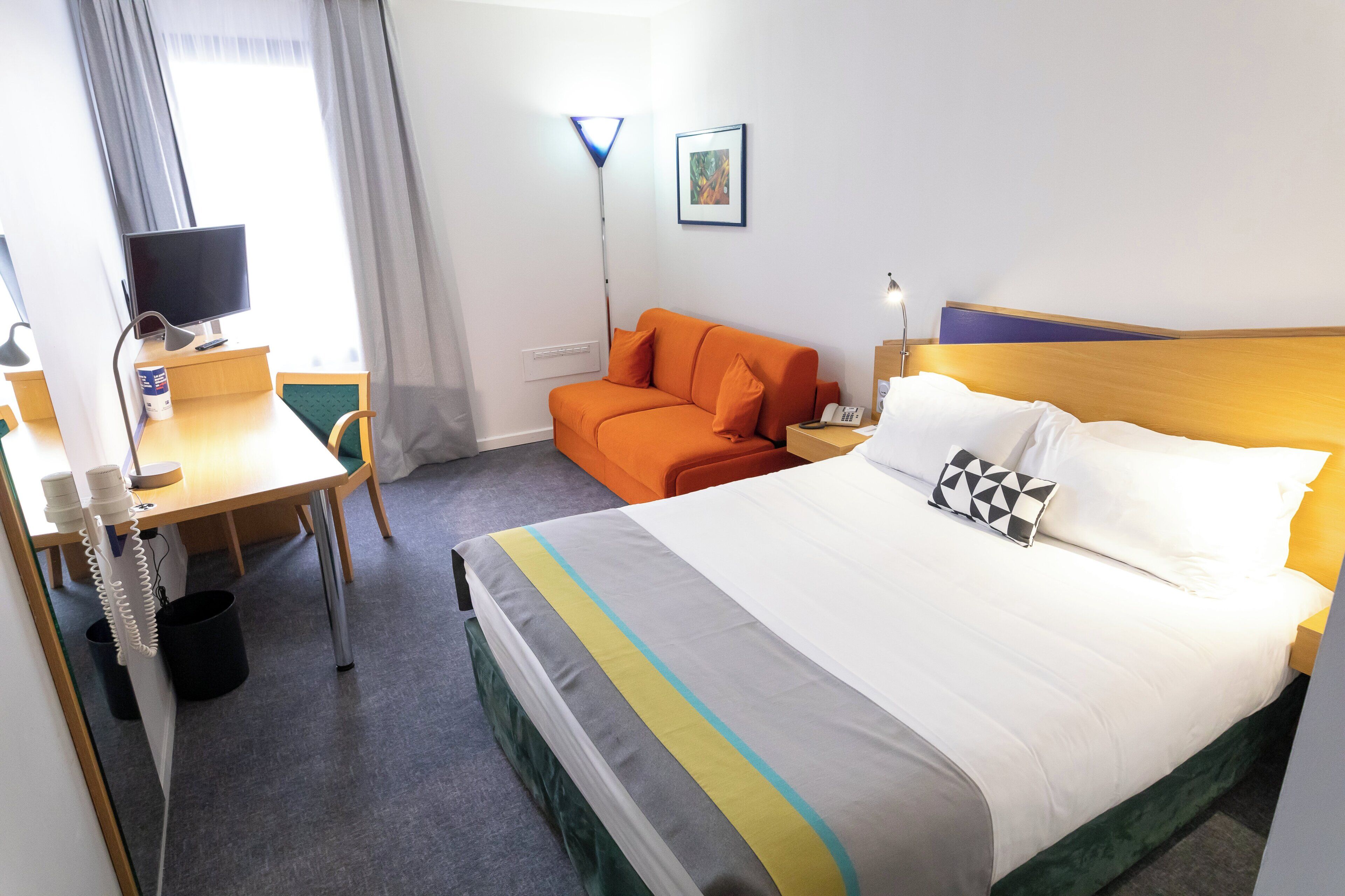A hotel room with a bed, an orange sofa, a desk with a computer, and a TV.