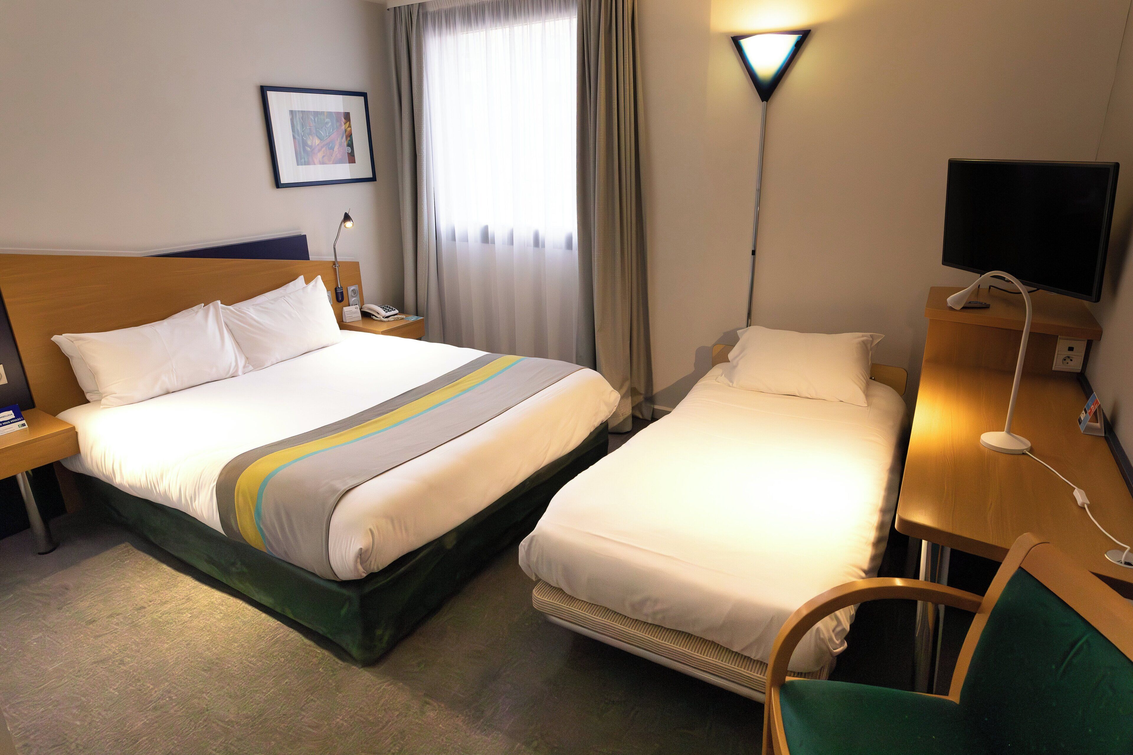 A hotel room with a double bed, a single bed, a desk with a chair, a TV, and a lamp.