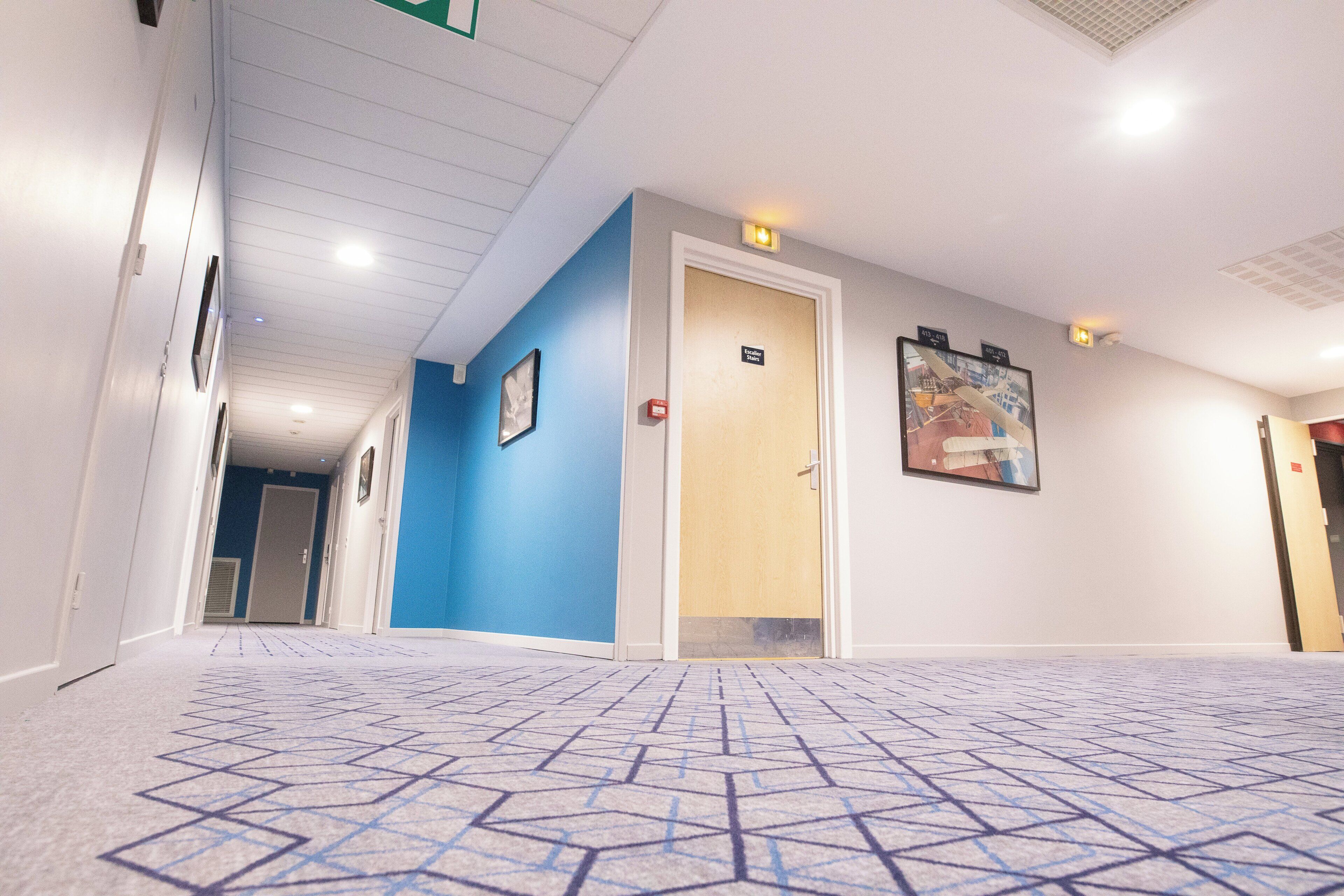 Un couloir d’hôtel avec un tapis à motifs géométriques, un mur bleu et une porte portant l’inscription « Escalators ».