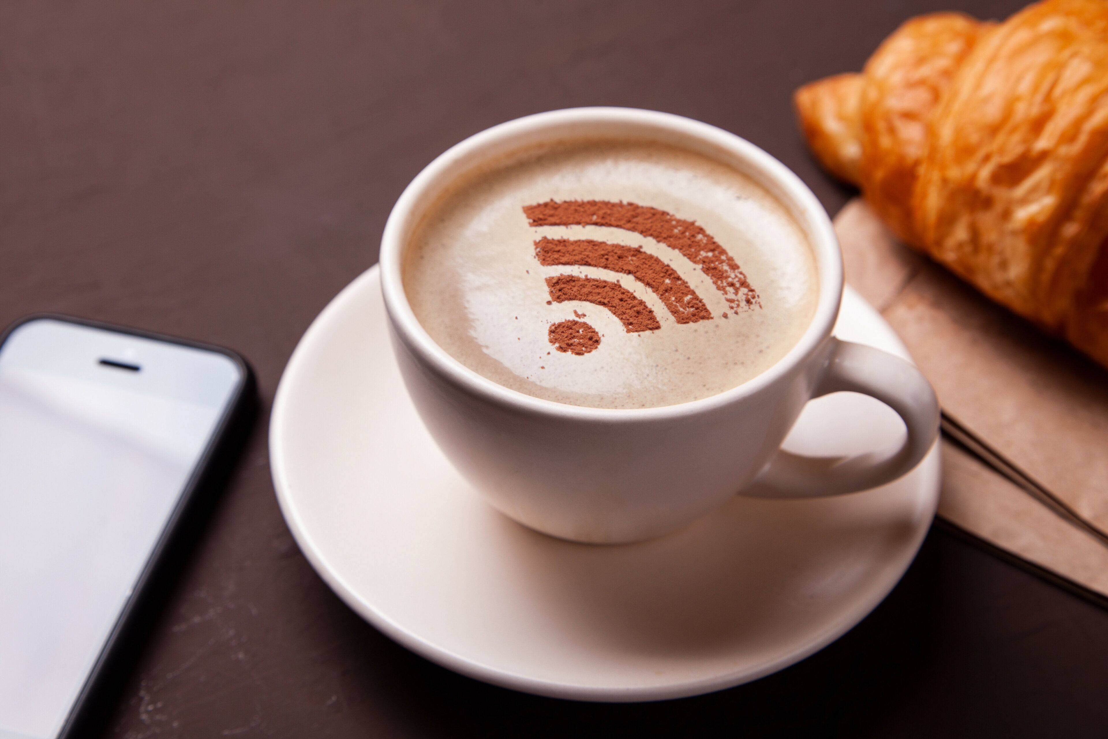 Una taza de café con un símbolo de Wi-Fi en la espuma, un croissant y un teléfono inteligente sobre una mesa.