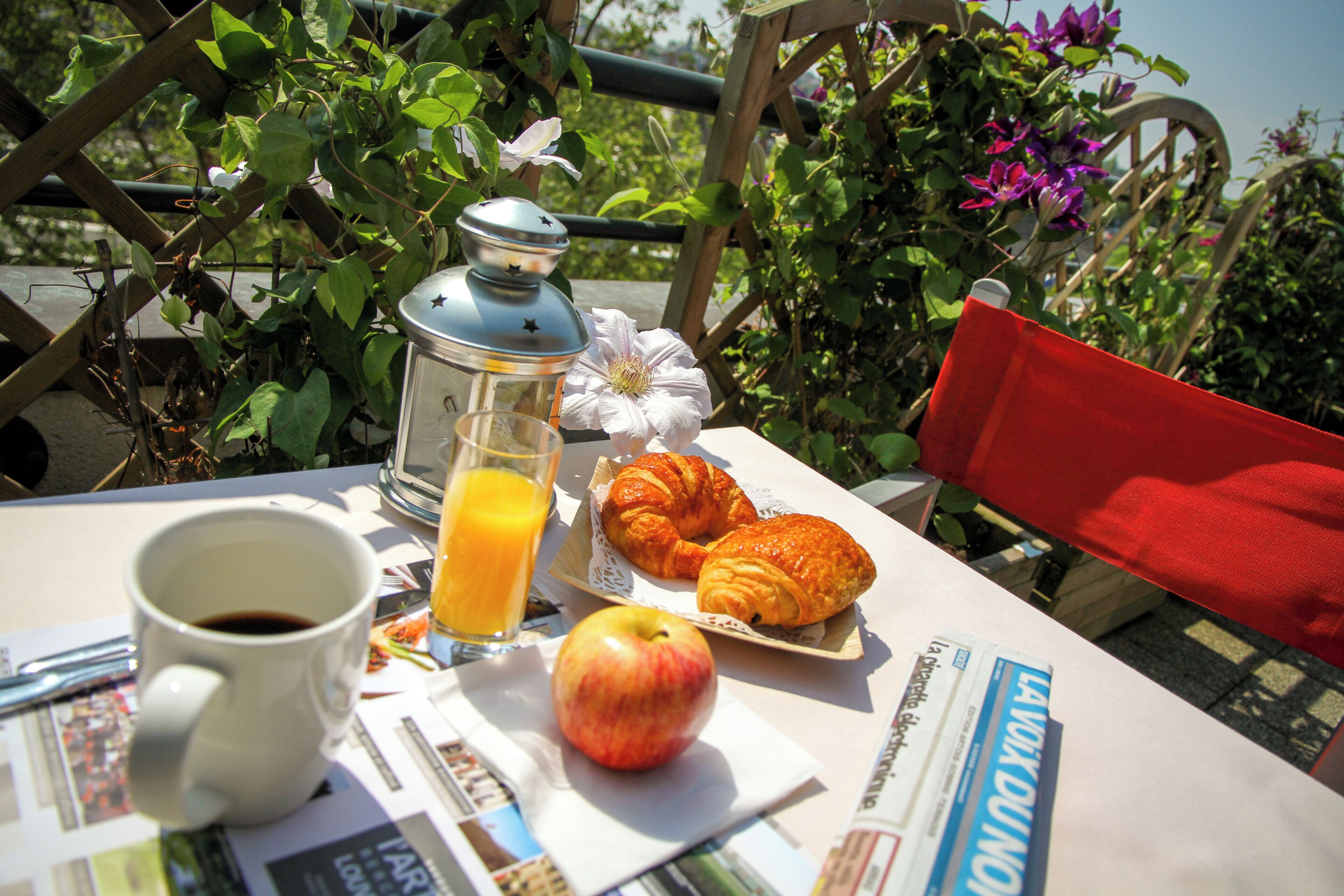Una mesa con una taza de café, un vaso de jugo de naranja, una manzana y croissants, ubicada al aire libre con follaje y una silla roja al fondo.