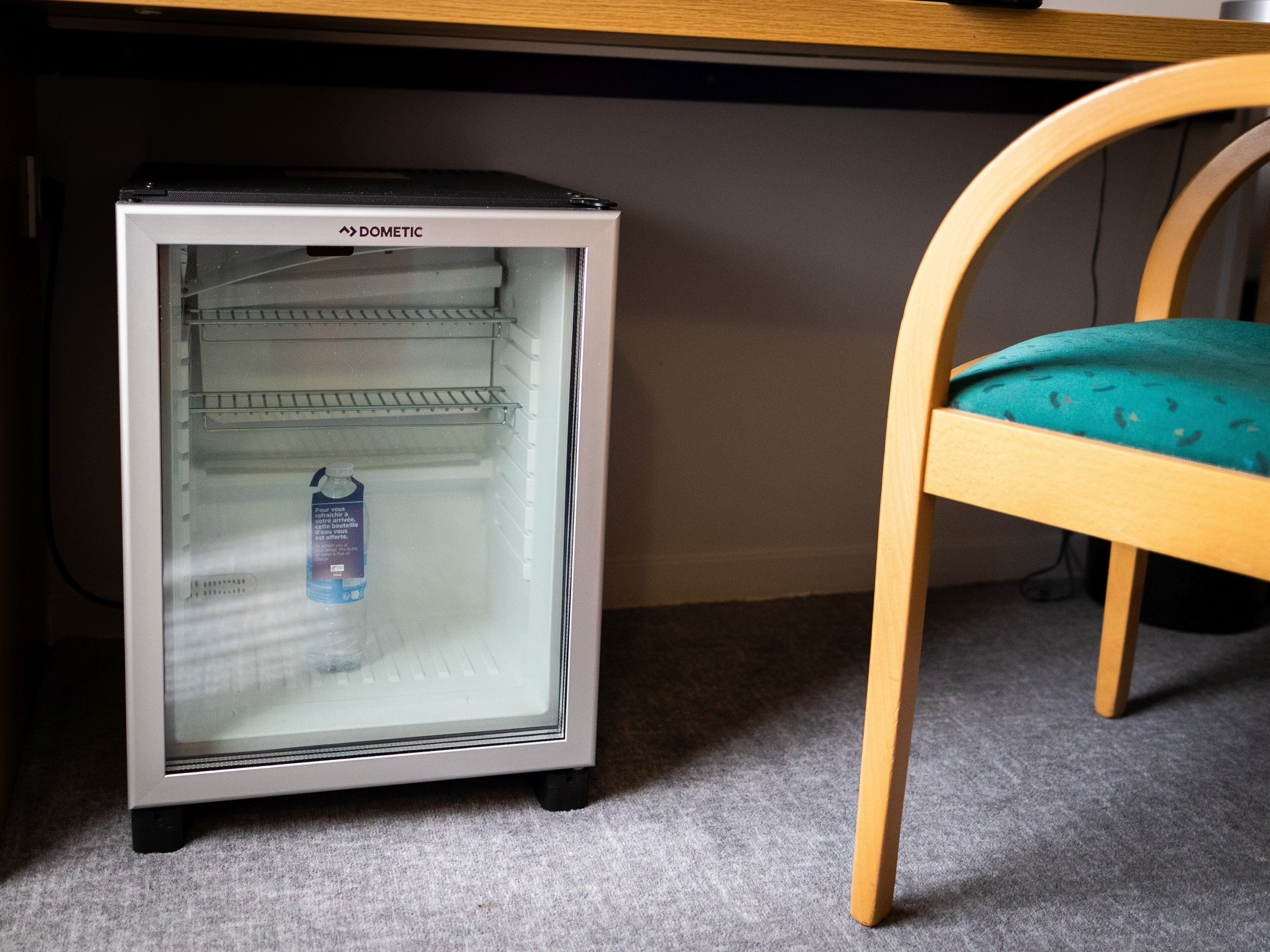 Ein kleiner, metallischer Kühlschrank mit Glastür und einer einzelnen Flasche im Inneren, steht auf einem Teppich neben einem Holzstuhl mit einem türkisfarbenen Kissen.