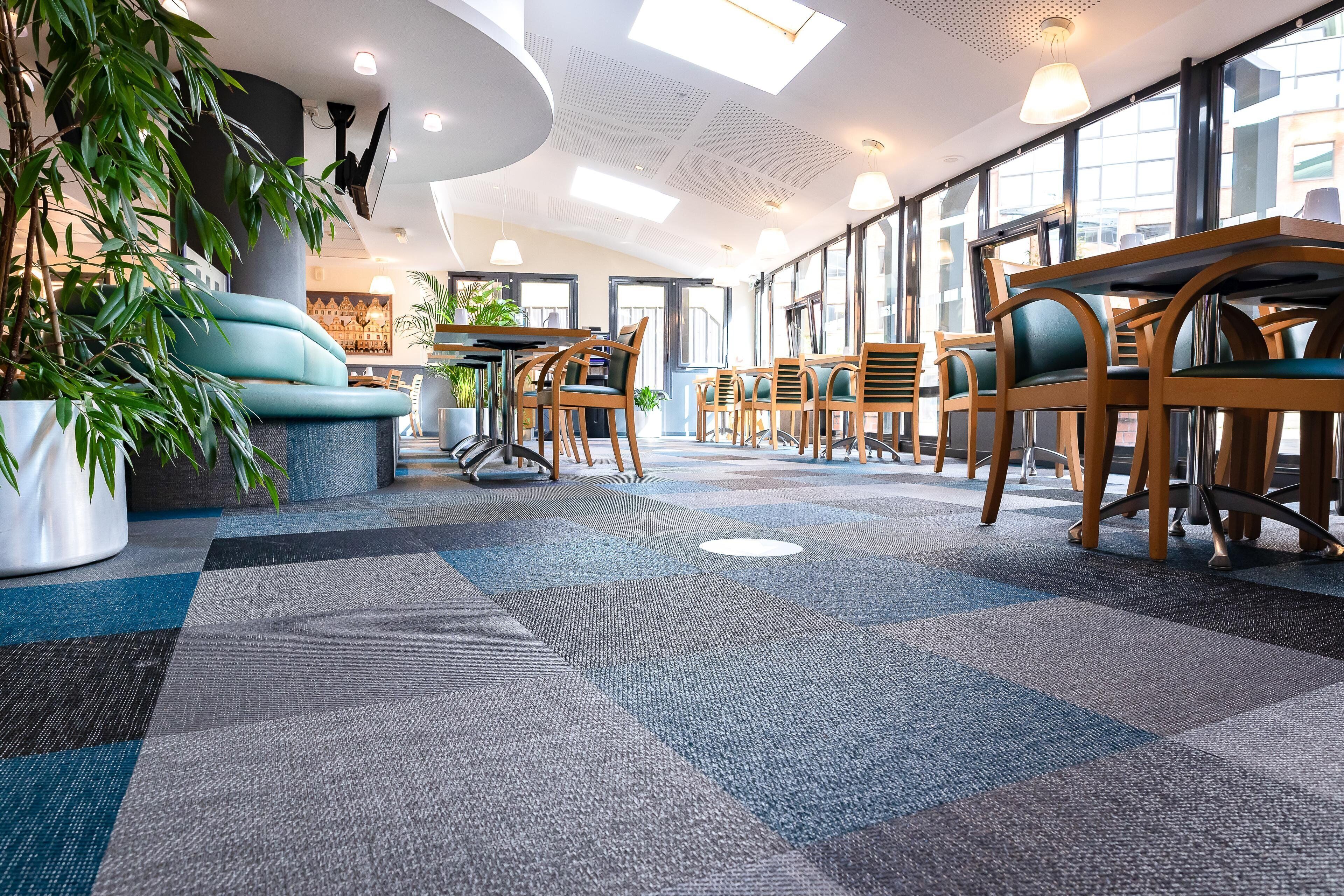 Eine moderne Hotellobby mit verschiedenen Sitzgelegenheiten, darunter eine Couch und mehrere Tische mit Stühlen. Üchtzipflanzen und ein großes Fenster sorgen für natürliches Licht.